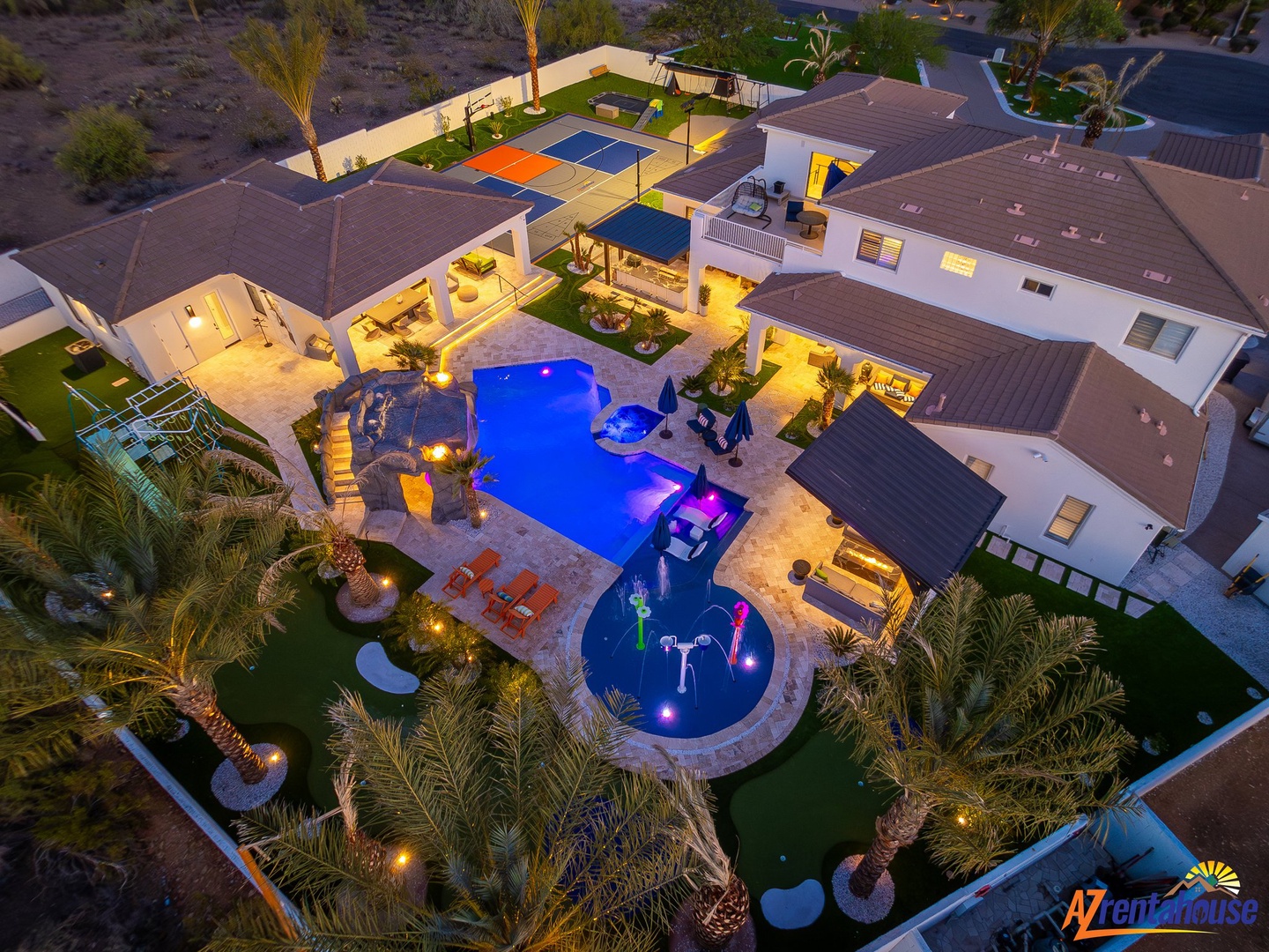 Stunning aerial view of this luxury vacation home featuring a spectacular pool with spa, sports court, and beautifully landscaped grounds illuminated at twilight.