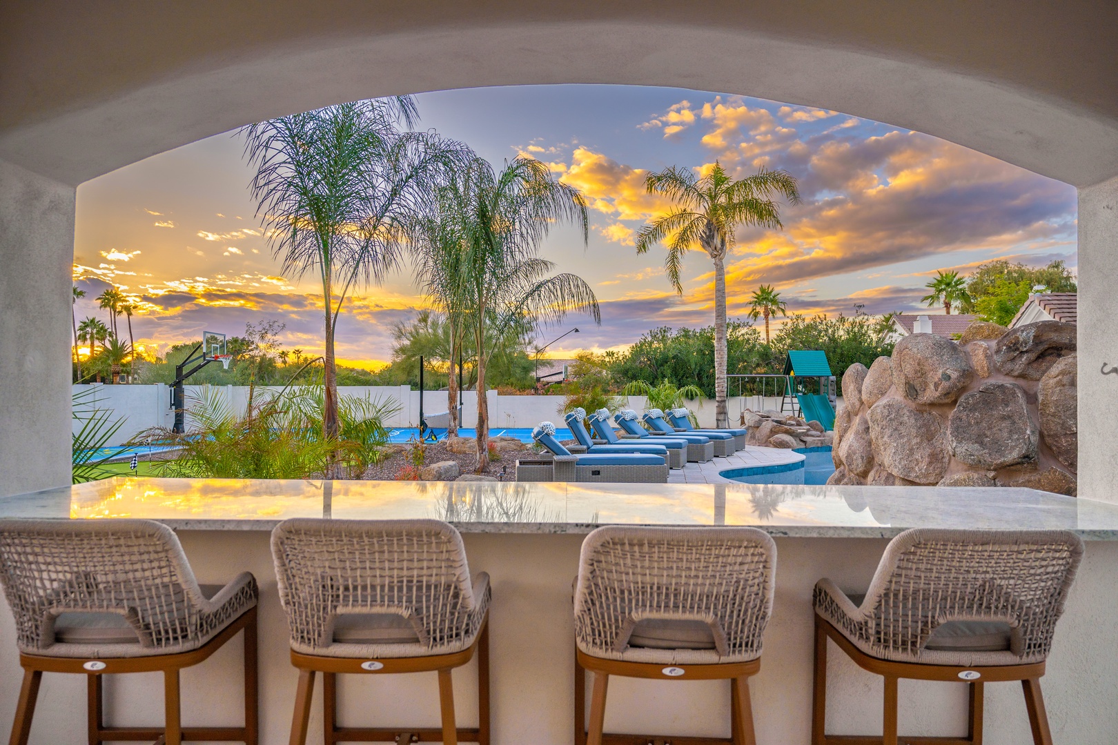 A stunning property framed by palm trees and dramatic golden-hour skies, featuring an outdoor pool, rock features, and lounge seating in a lush desert oasis setting.