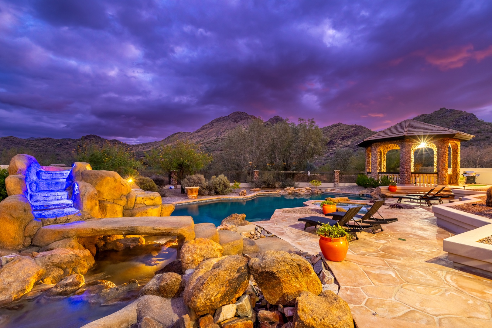A stunning desert mountain backdrop frames this resort-style backyard with illuminated pool, cascading rock waterfall with blue lighting, and covered gazebo at twilight.
