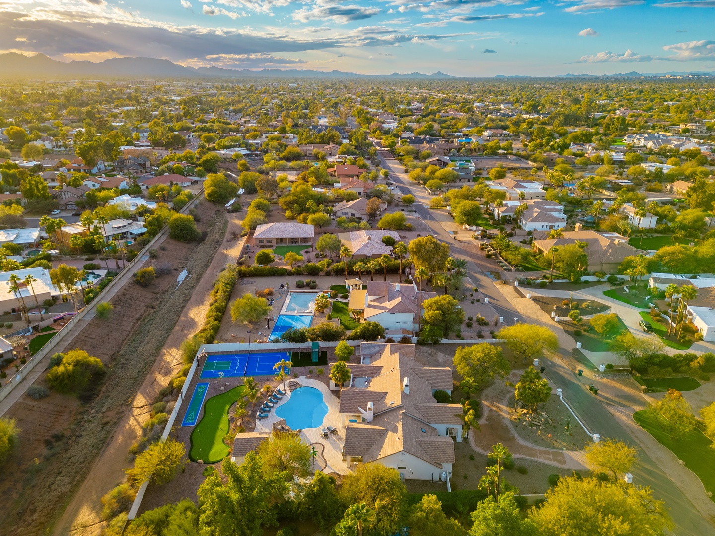 Aerial view of the property nestled in a scenic neighborhood, featuring two swimming pools, sports courts, and lush desert landscaping with mountain views on the horizon.