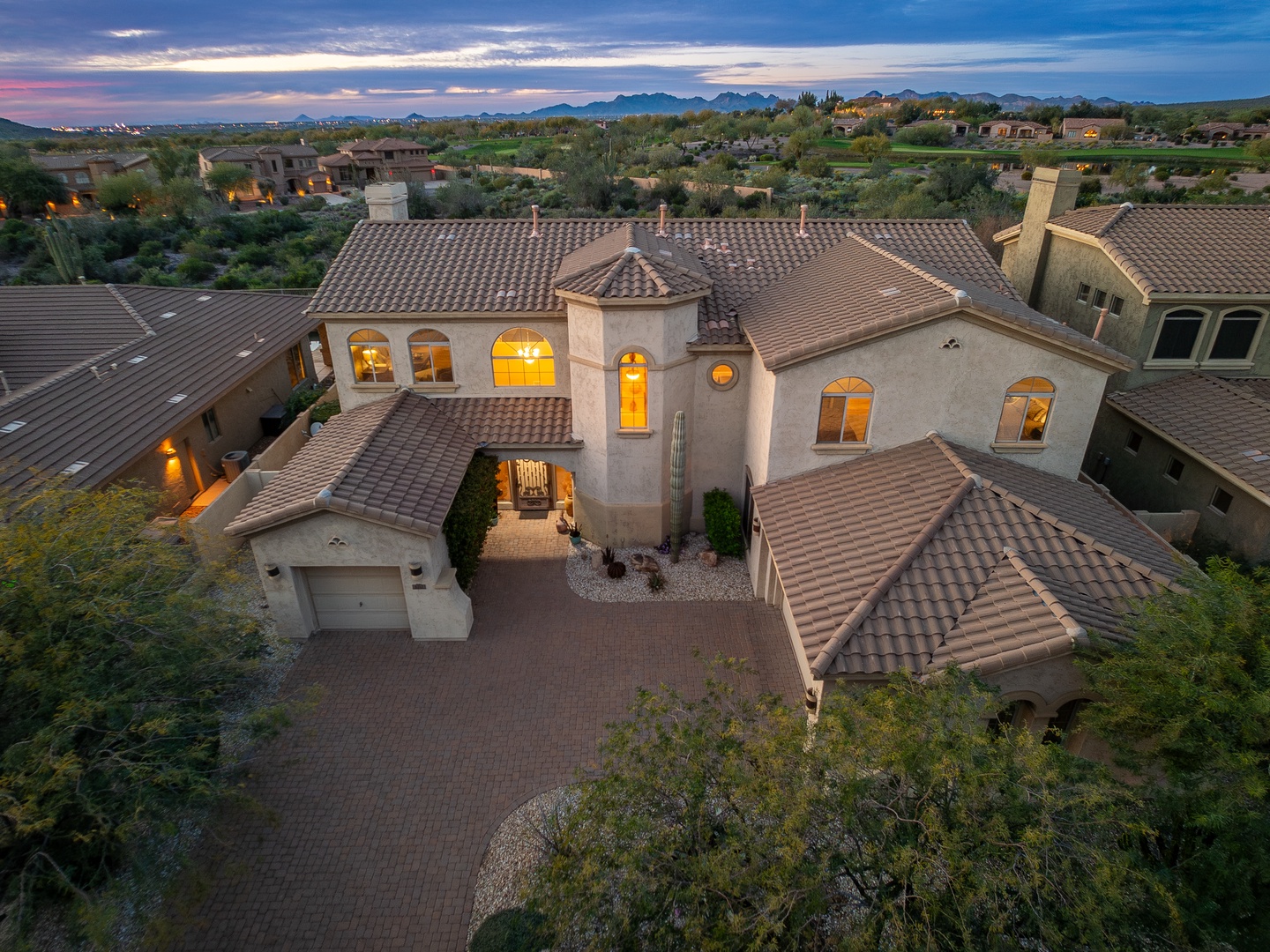 Aerial view of an elegant desert home featuring Spanish-style architecture, surrounded by lush landscaping and mountain vistas. The property sits within a serene neighborhood, offering tranquil desert charm.
