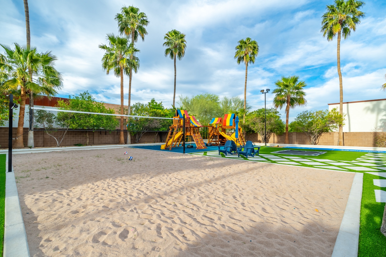A spacious outdoor play area with swings, slides, and a sand volleyball court, all set under swaying palm trees and sunny skies.
