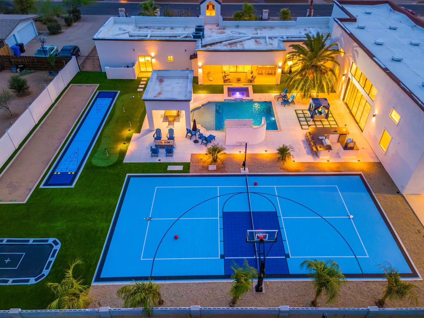 Aerial view of a luxury vacation property showcasing illuminated outdoor entertainment spaces with a full-size sport court, lap pool, main pool, patio dining areas, and tropical landscaping at dusk.
