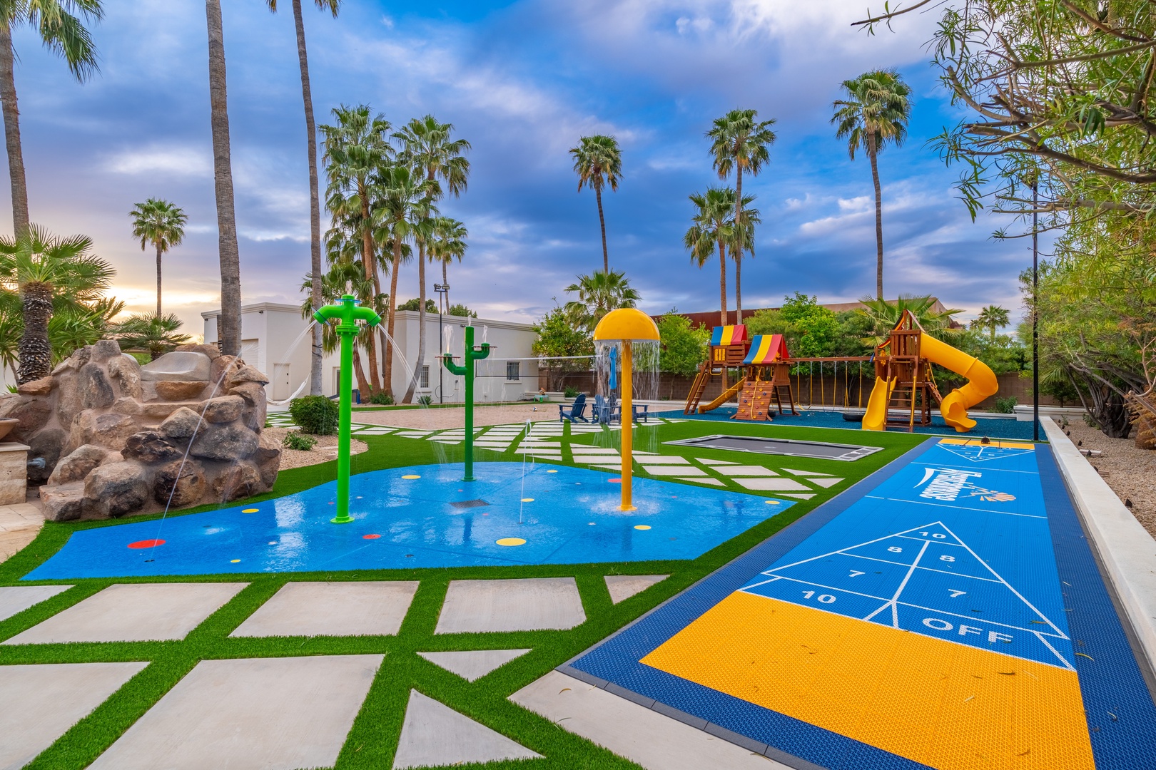 A vibrant outdoor play area featuring a splash pad, playground equipment, and interactive games beneath swaying palm trees at dusk.