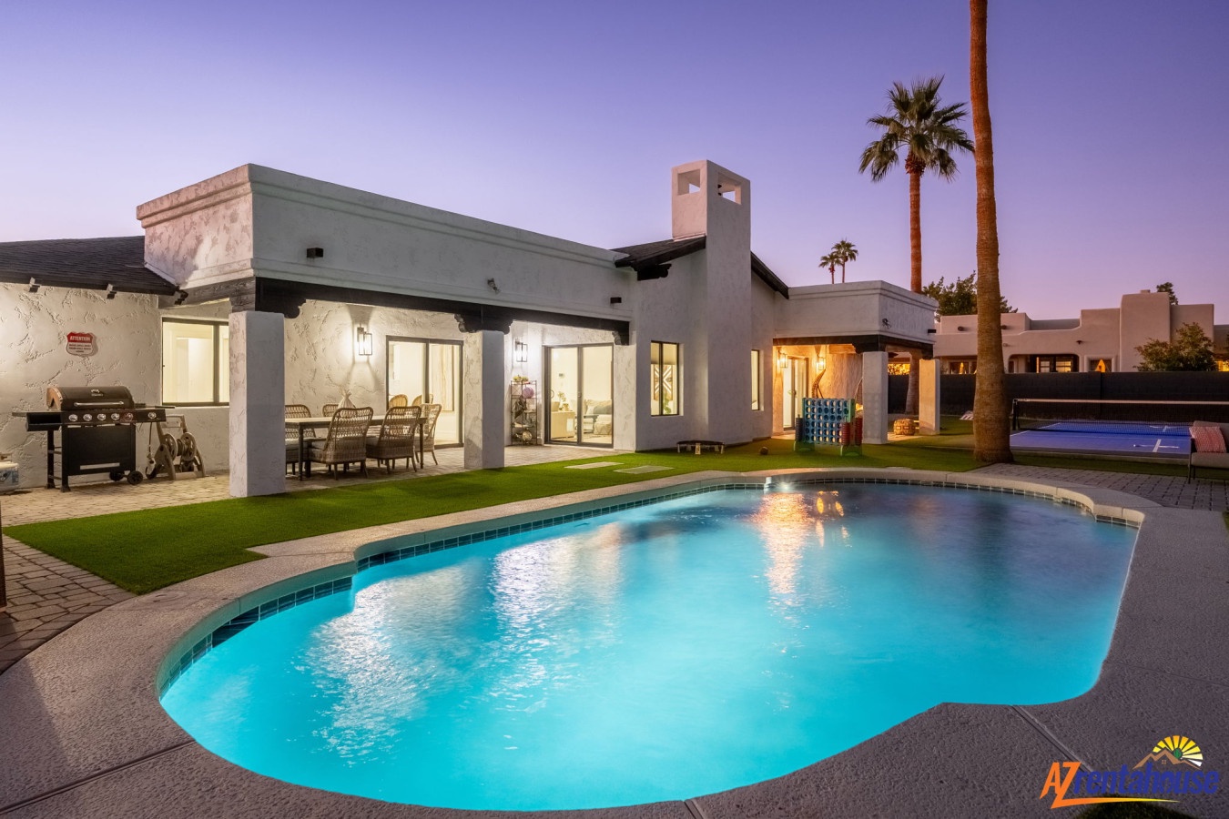 Modern vacation home features a pristine swimming pool, outdoor dining, and BBQ area under desert palms at twilight.