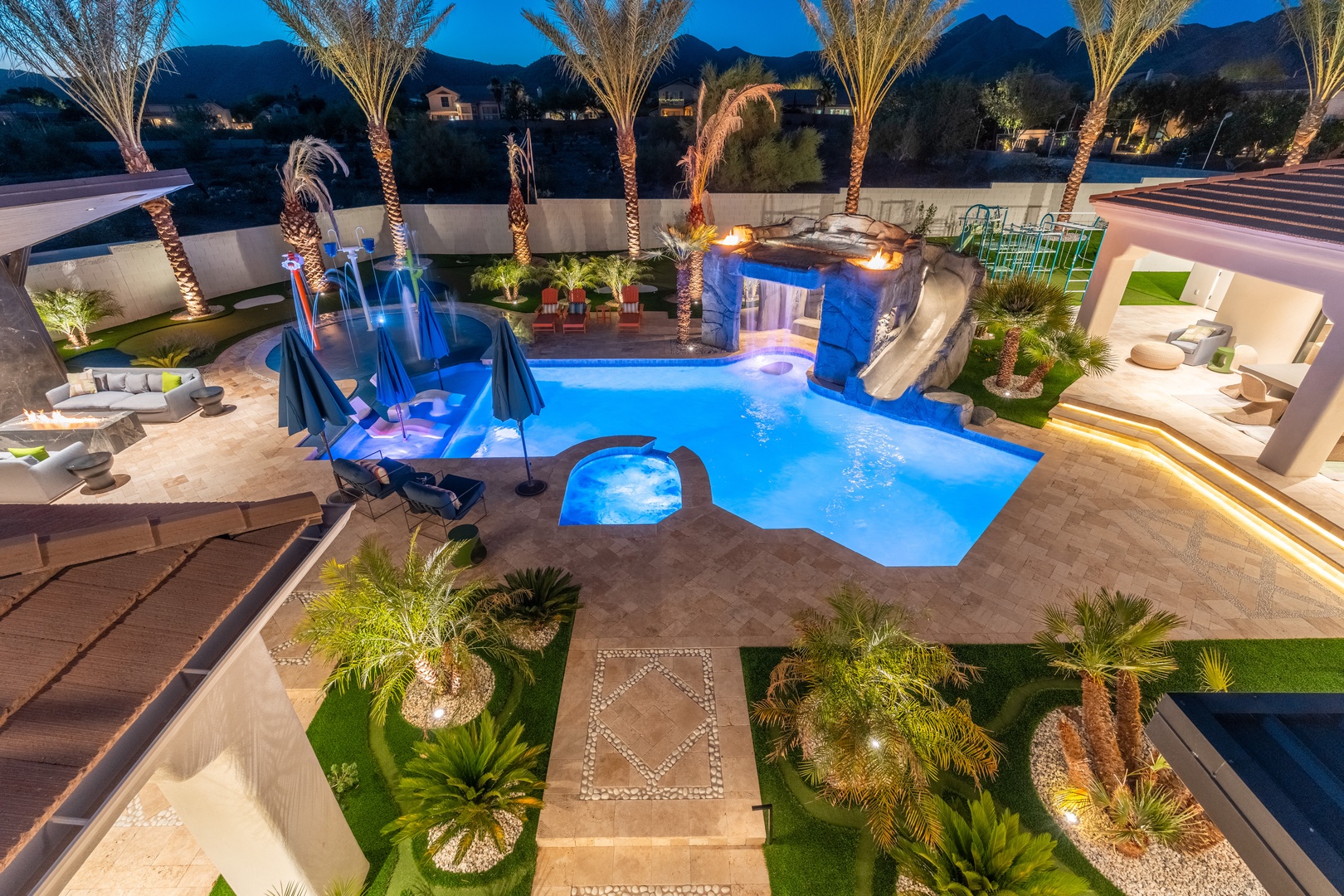 An aerial view captures the stunning backyard resort area, featuring a dramatic free-form pool with water slides, fire features, and illuminated palm trees set against a mountainous desert backdrop at dusk.