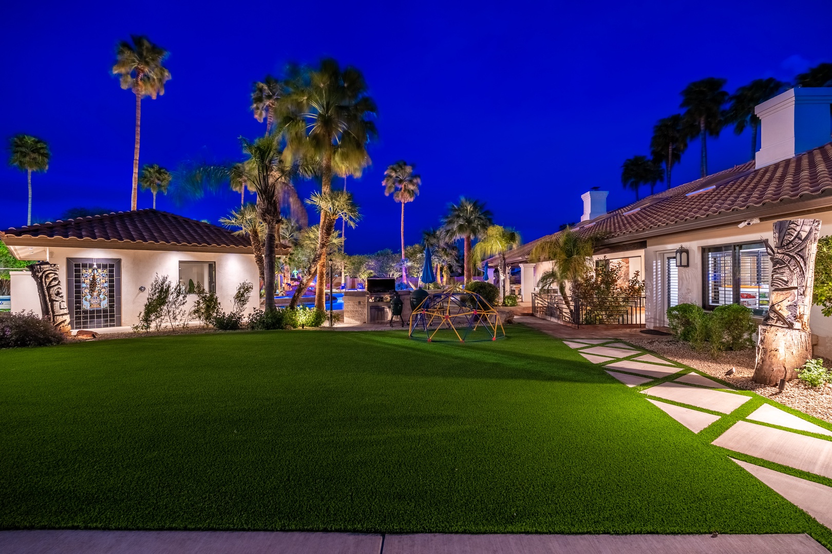 A stunning property exterior showcasing Spanish-style villas with red tile roofs, tropical palm trees, and manicured lawns under a dramatic twilight sky, with a playground area illuminated in the background.