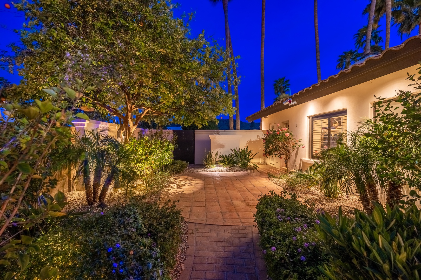 The property's entrance pathway showcases Spanish-style architecture with warm lighting, mature trees, and lush desert landscaping featuring palms and flowering plants, creating a welcoming resort-like atmosphere.
