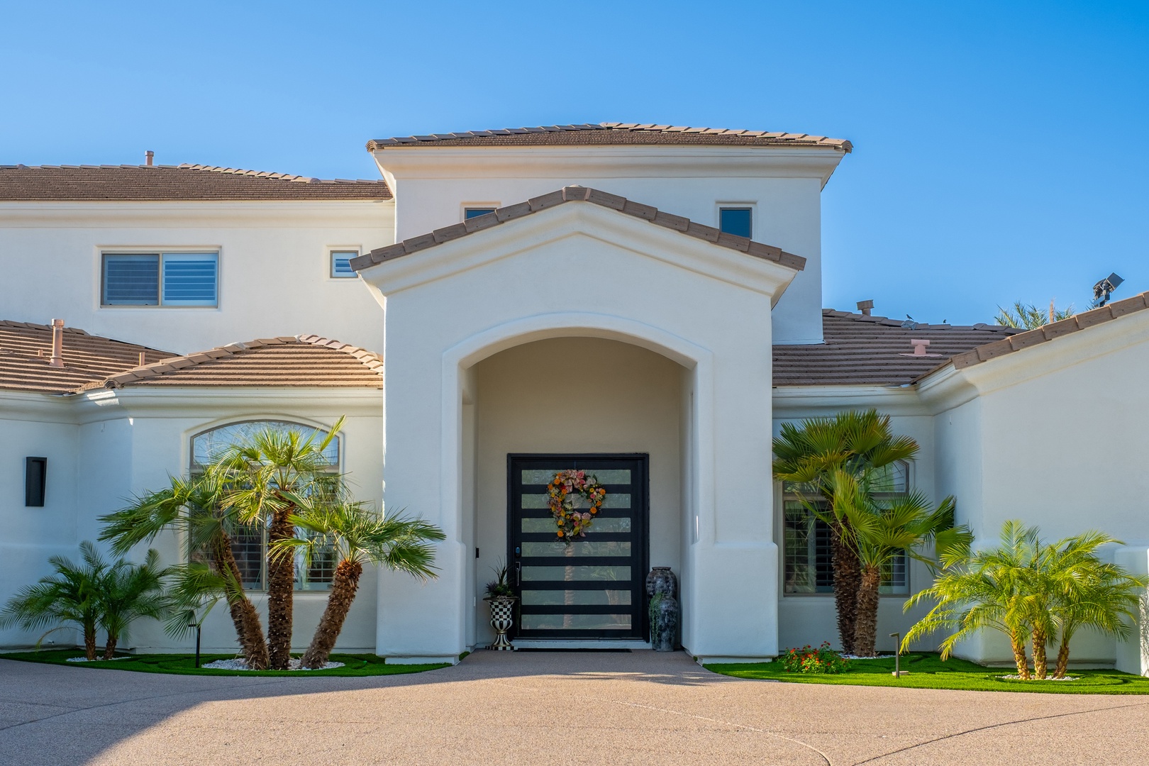 A striking modern home with elegant white stucco architecture, arched entryway, and lush palm landscaping under clear blue skies.