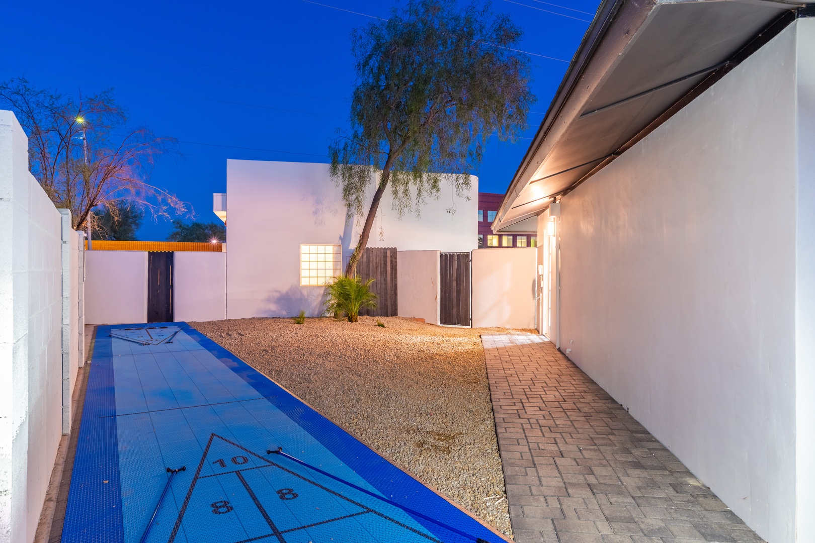 Modern courtyard with lap pool and gravel landscaping under desert twilight. Clean white walls and paver walkway lead to illuminated entrance and private outdoor spaces beyond.