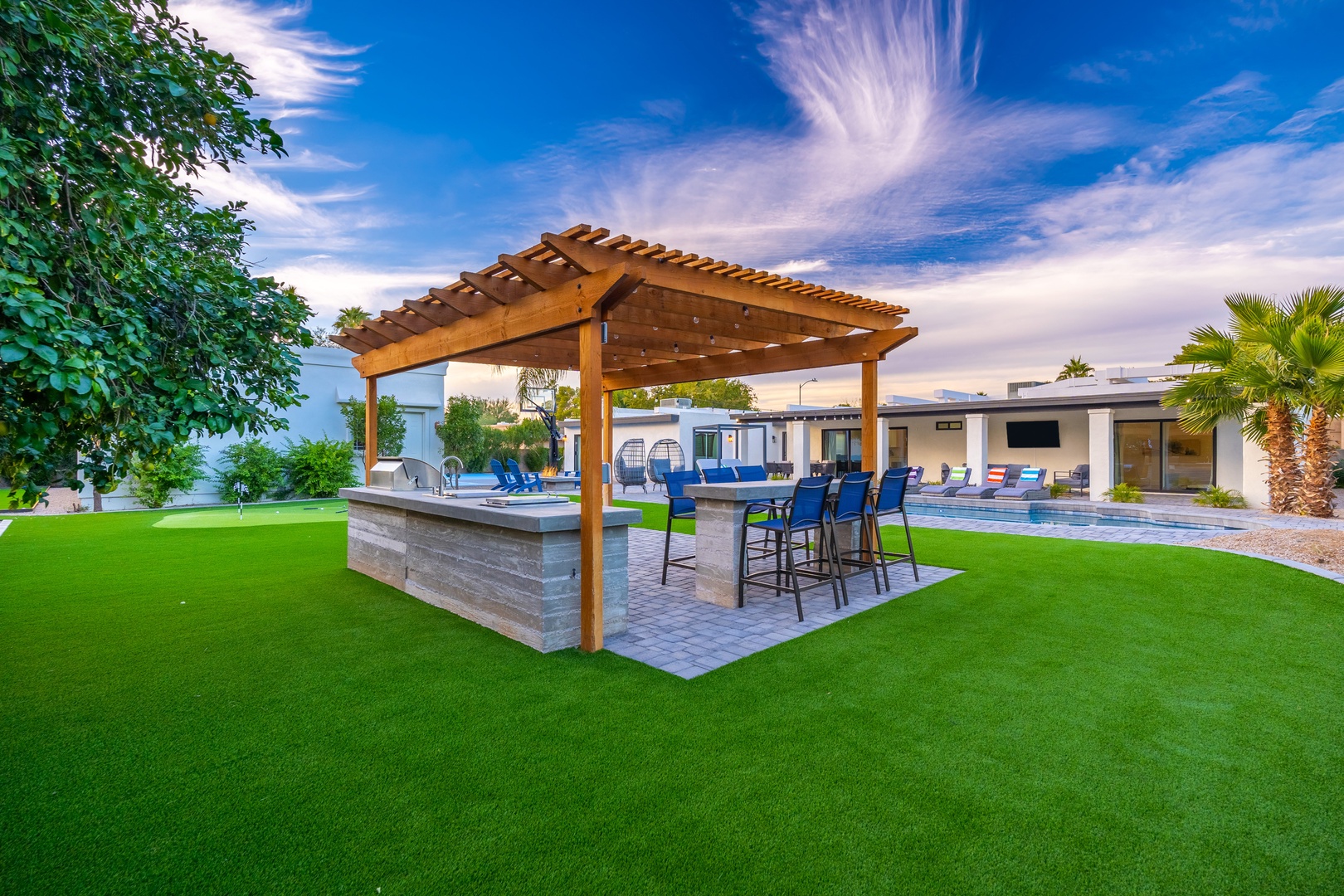 A stunning outdoor oasis featuring an expansive lawn, a modern pergola-covered bar, a sparkling pool, and lush landscaping under a dramatic sky—perfect for outdoor entertaining and relaxation.