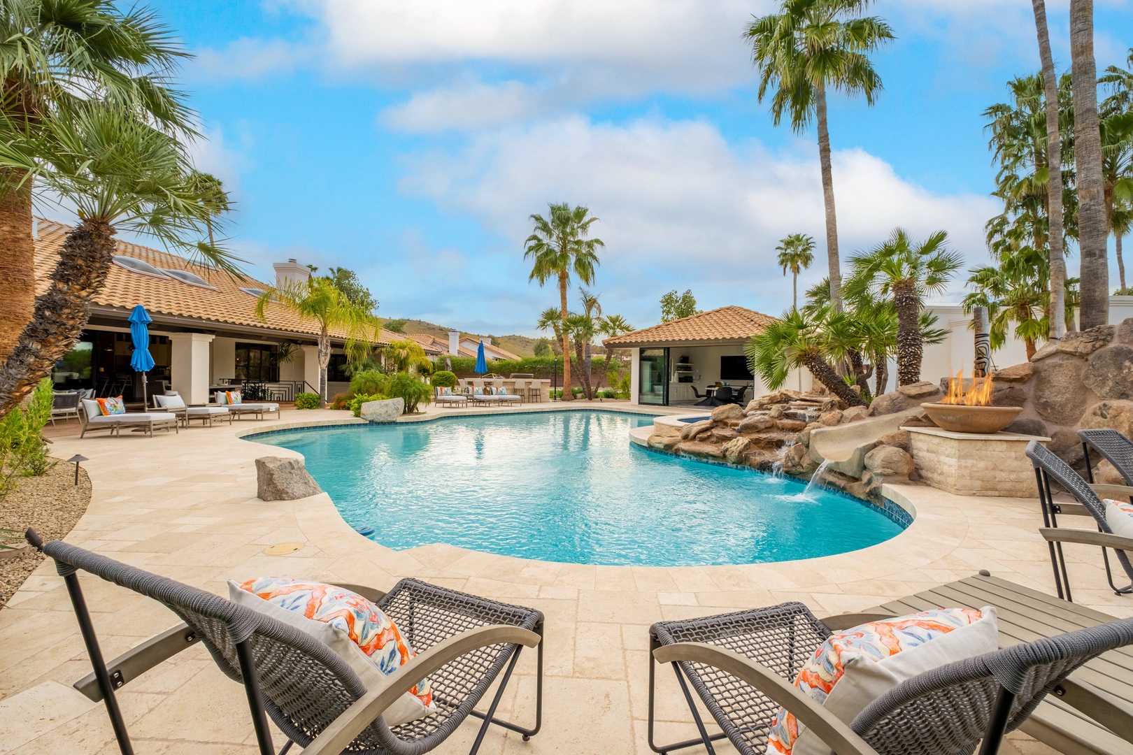 A stunning resort-style pool with rock waterfall features awaits, framed by swaying palms and inviting sun loungers beneath clear desert skies.