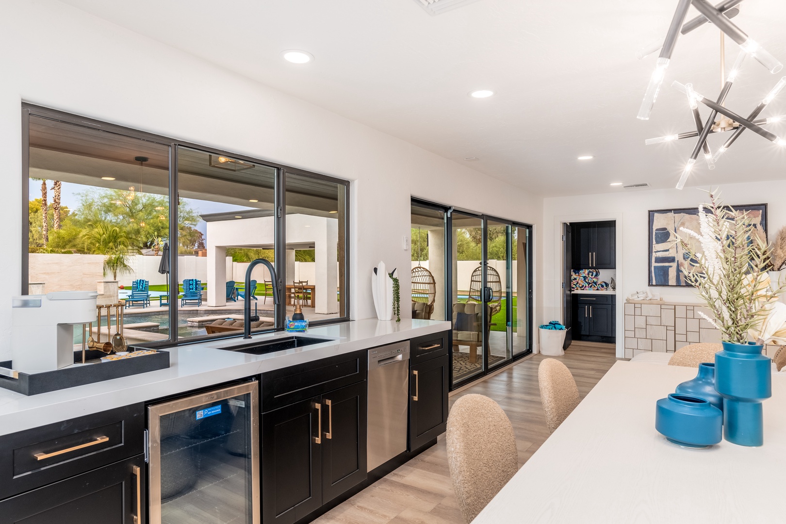 Your sleek kitchen opens to a sun-soaked patio with floor-to-ceiling glass doors. Modern black cabinetry contrasts with white countertops, while blue accent pieces add a welcoming pop of color throughout.
