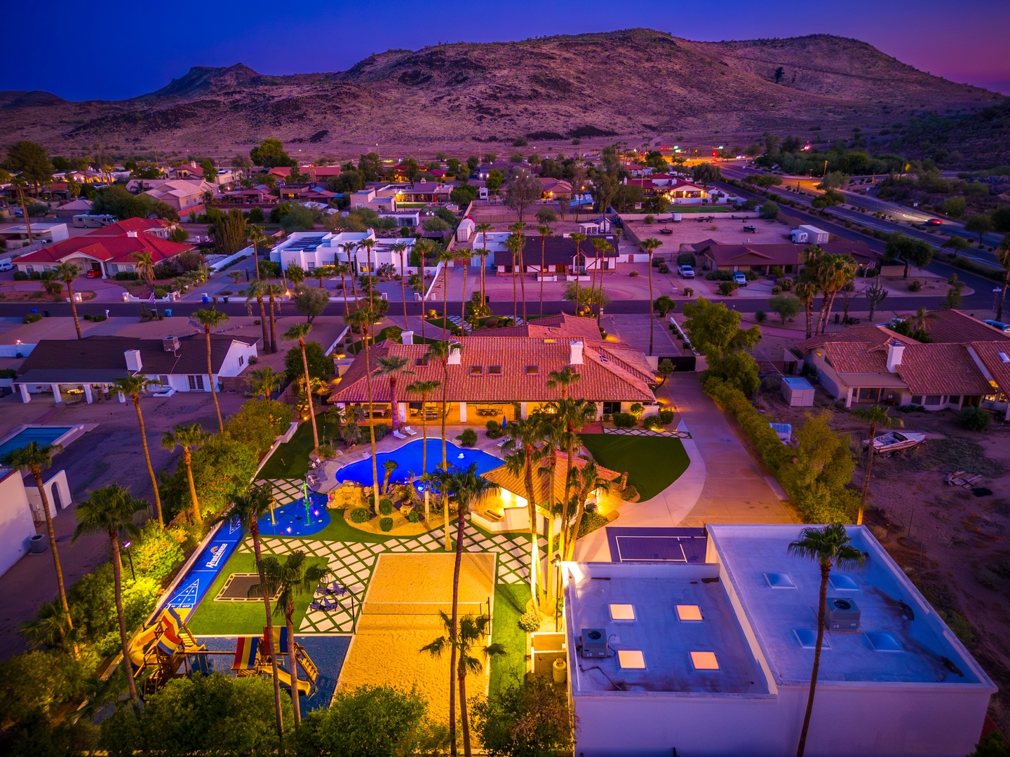 Aerial view of a desert vacation property at twilight, featuring an illuminated pool, lush palm gardens, and mountain backdrop in a peaceful residential setting.