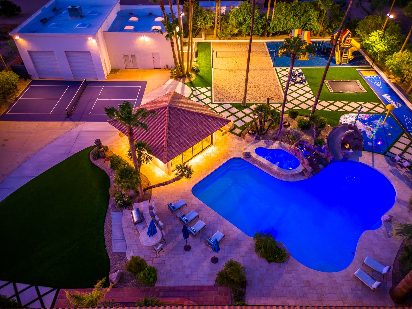 Aerial view of an expansive vacation rental estate showcasing a striking illuminated pool, private tennis court, hot tub, playground, and lush tropical landscaping—ideal for families and groups seeking luxury and recreation.