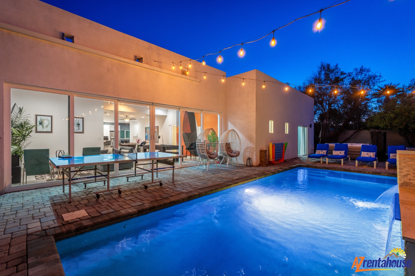 Evening pool illuminated in brilliant blue creates magical backyard ambiance with string lights overhead and modern patio seating.