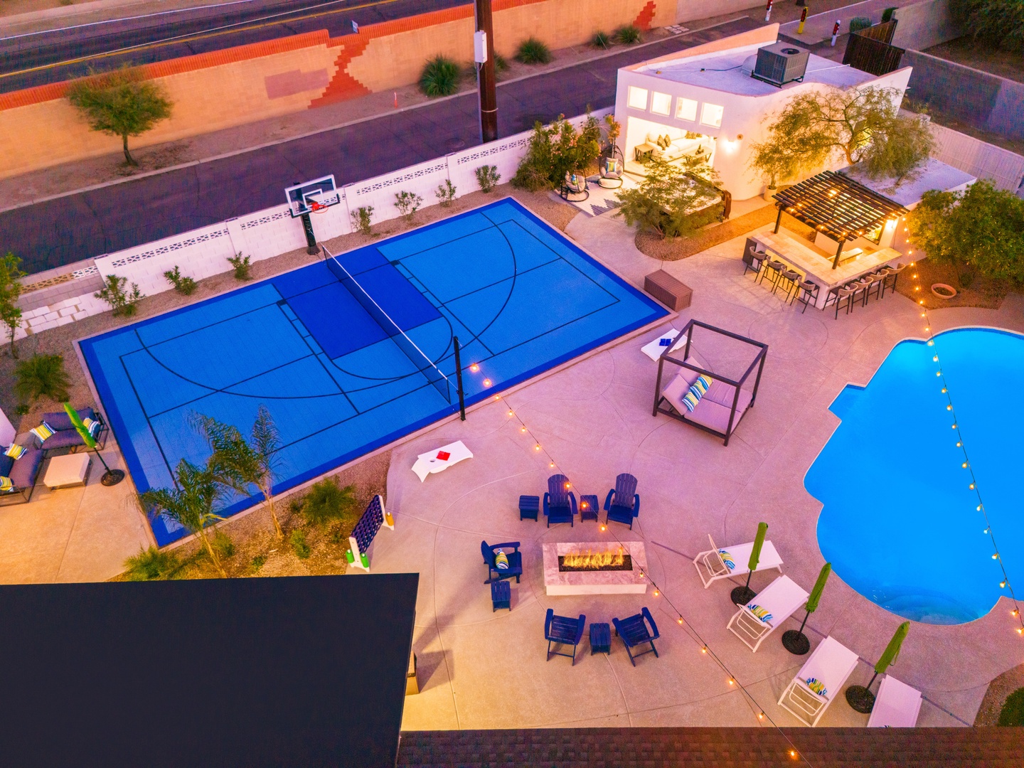 Aerial view showcasing an expansive backyard featuring a sports court, swimming pool, outdoor dining area with pergola, fire pit lounge, and mature landscaping—highlighting the property's extensive recreational amenities and gathering spaces.