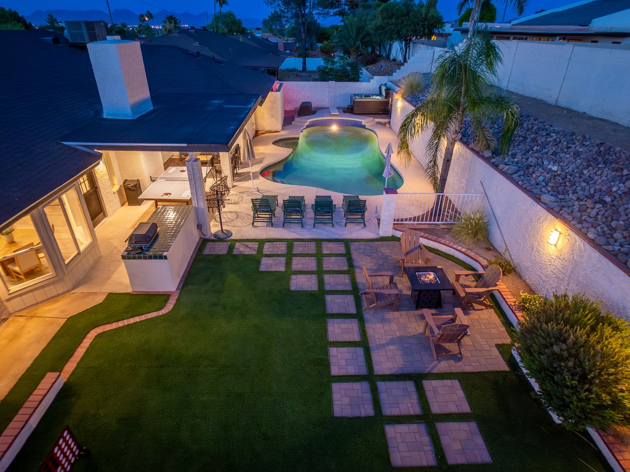 Modern vacation home featuring illuminated pool, hot tub, and multiple outdoor entertaining areas set against a twilight sky with palm trees and mountain views in the surrounding neighborhood.