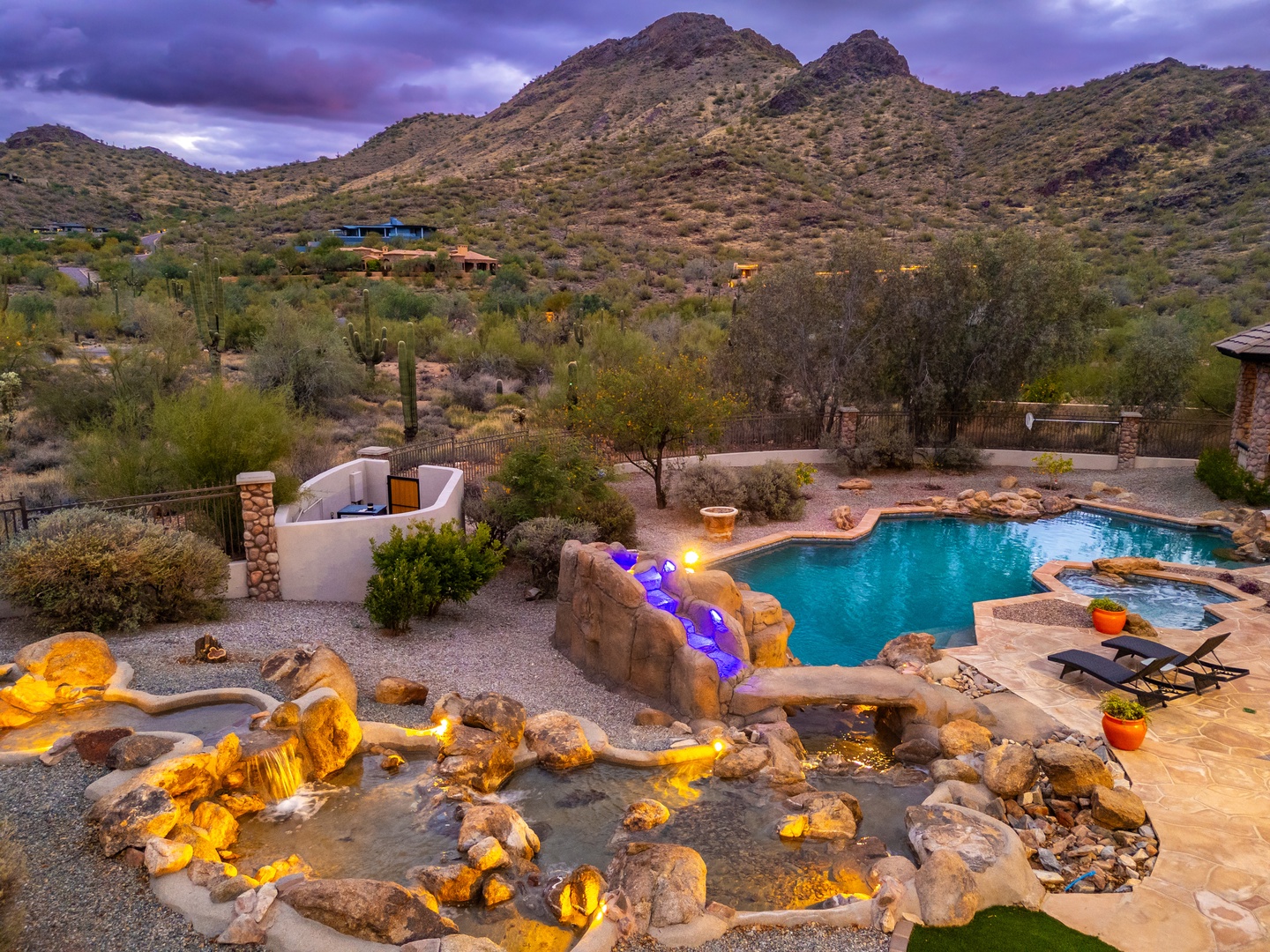 A stunning desert mountain setting frames this private oasis with pool, hot tub, and illuminated waterfall feature—serene luxury amid rugged natural beauty at twilight.