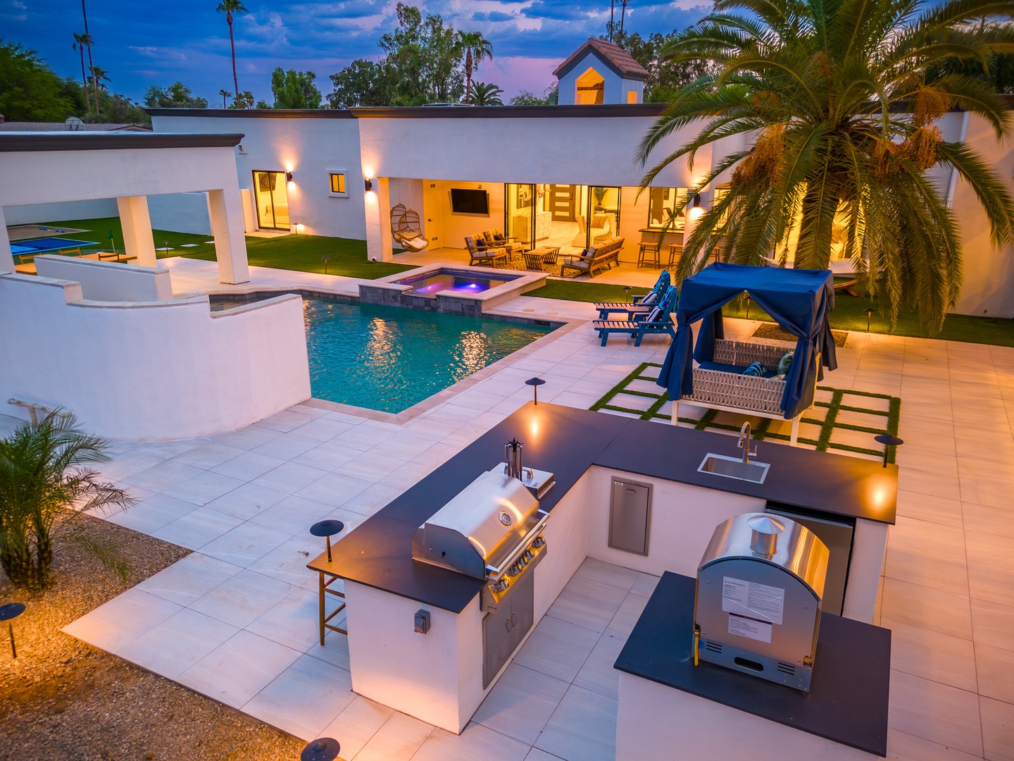 This modern property features a sparkling pool, spa, outdoor kitchen with BBQ facilities, and covered lounge areas, all framed by palm trees and warm evening lighting in a serene neighborhood setting.