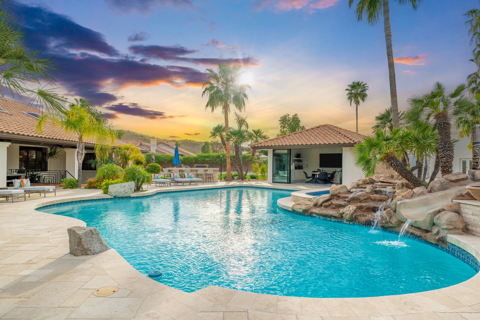 A stunning resort-style pool oasis framed by swaying palms and desert hills, featuring a cascading rock waterfall and spacious patio—perfect for unwinding under painted skies.