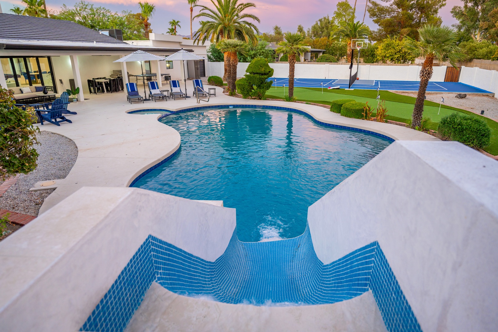 This beautifully landscaped backyard oasis features a sparkling pool, covered patio dining, a tennis court, and lush tropical gardens surrounded by palm trees under a stunning desert sunset.