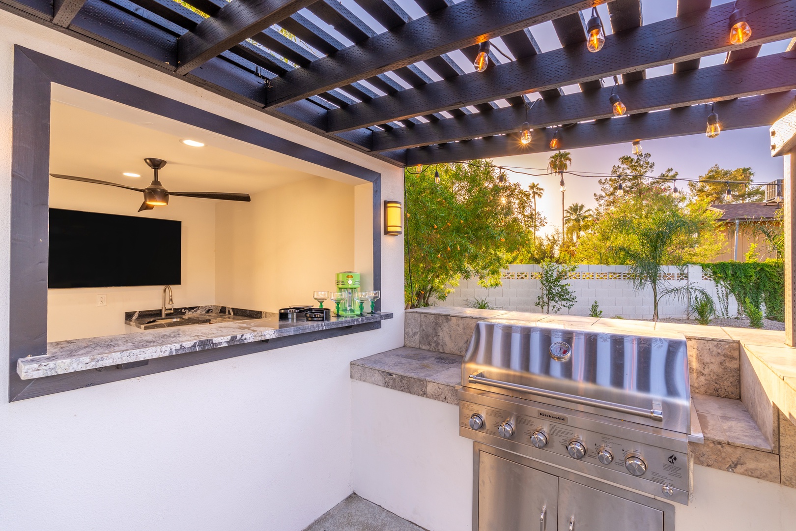 Your private outdoor kitchen awaits with a gleaming stainless steel grill, granite counters, and built-in TV—all under a stunning pergola with ambient string lights perfect for evening entertaining.