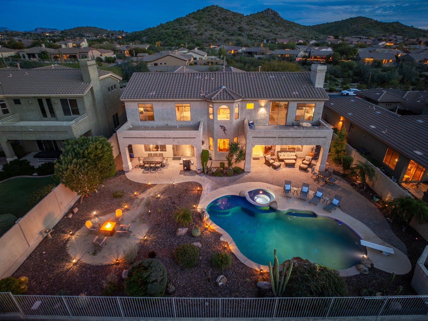 Aerial view of a luxurious desert retreat featuring a sparkling pool, hot tub, and expansive outdoor entertaining spaces set against scenic mountain foothills at twilight.