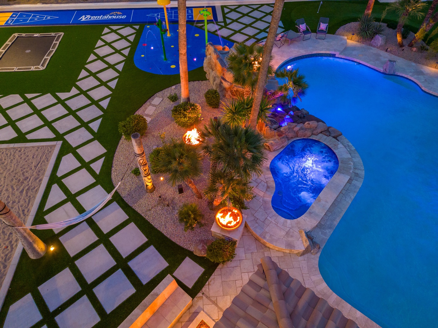 Aerial view of a resort-style backyard featuring a large swimming pool, hot tub, sport courts, manicured landscaping with palm trees, fire features, and outdoor seating areas illuminated at twilight.
