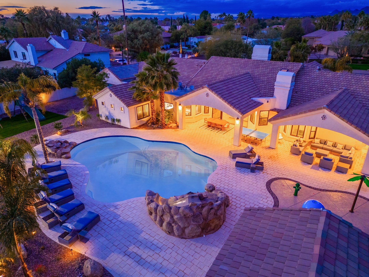Aerial view of the property at dusk, showcasing the expansive pool with integrated hot tub, spacious covered patio seating, and lush residential neighborhood surroundings.