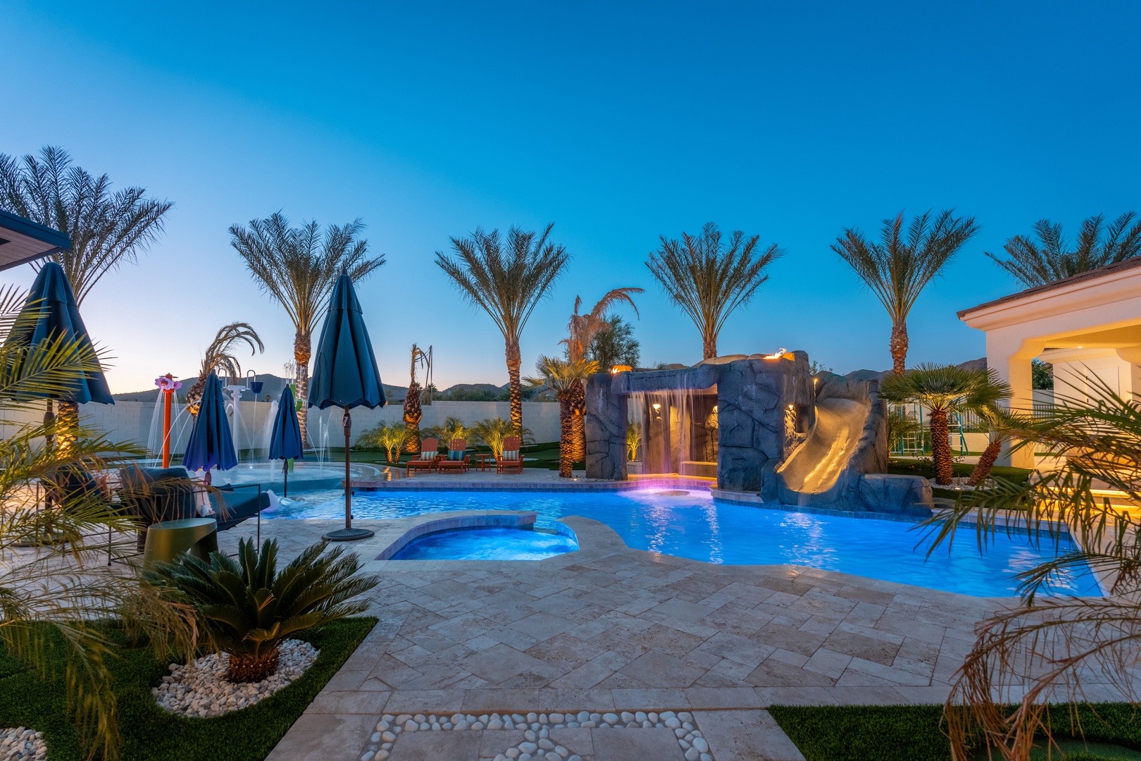 Splash into paradise! This sparkling pool features a thrilling rock waterslide, cozy hot tub, and tropical palms under desert skies—your perfect backyard oasis awaits.