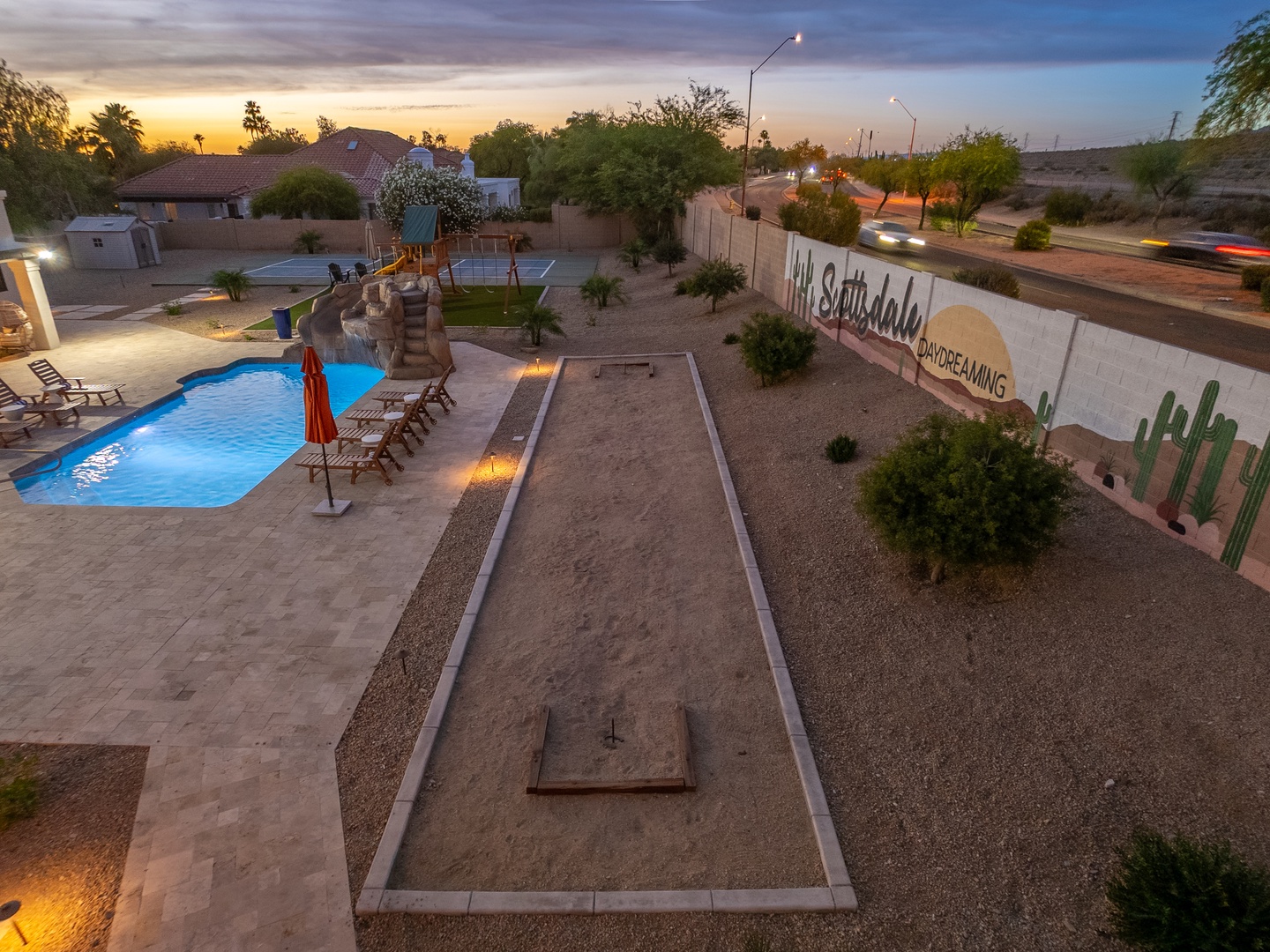 Aerial view of La Scottsdale vacation rental property at sunset, featuring a backyard with swimming pool, sport court, desert landscaping, and distinctive property signage along the street.