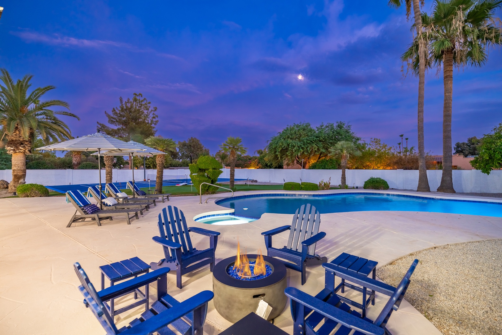 A twilight desert oasis featuring a sparkling pool, fire pit seating, and swaying palms beneath a moonlit sky—perfect for relaxed Arizona evenings.