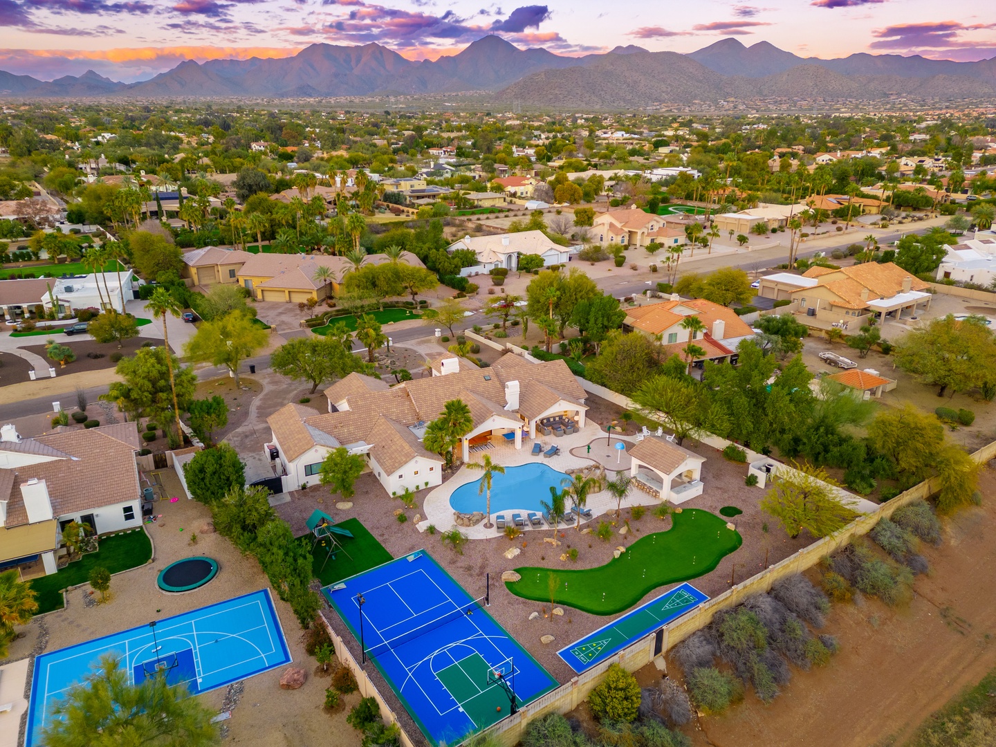 Aerial view of a desert vacation property showcasing tennis and sport courts, a blue swimming pool, and manicured lawn areas, set against a stunning mountain backdrop at sunset.