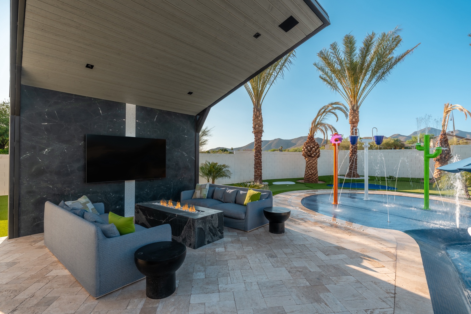 A covered patio combines comfort and entertainment with a sleek lounge, modern fireplace, and wall-mounted TV overlooking a vibrant splash pad and mountain views—perfect for family relaxation and play.