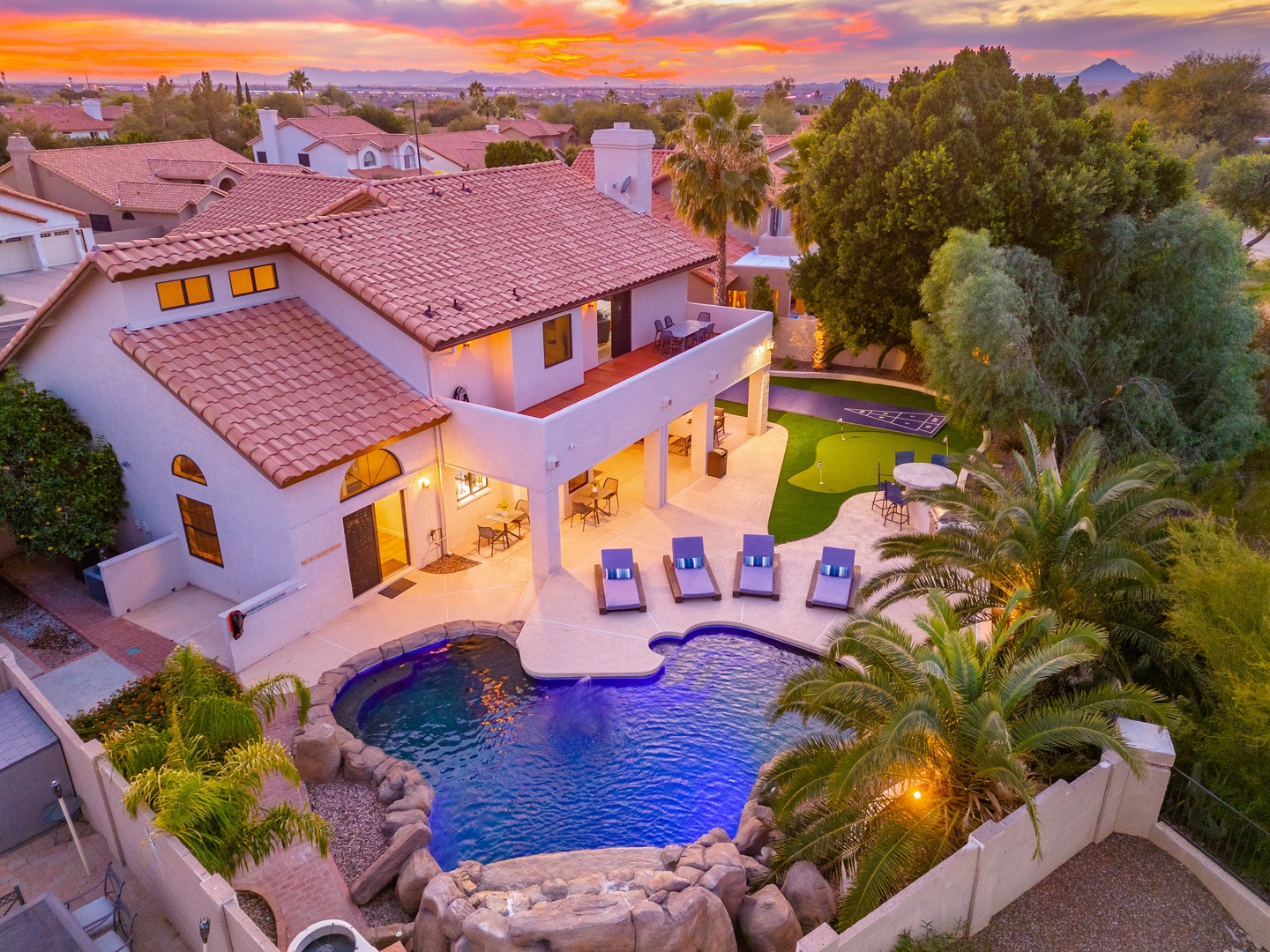Aerial view of a white stucco rental home with terra cotta tile roof, featuring a resort-style pool, spacious patio with lounge chairs, and lush desert landscaping, set against mountains and a vibrant sunset sky.