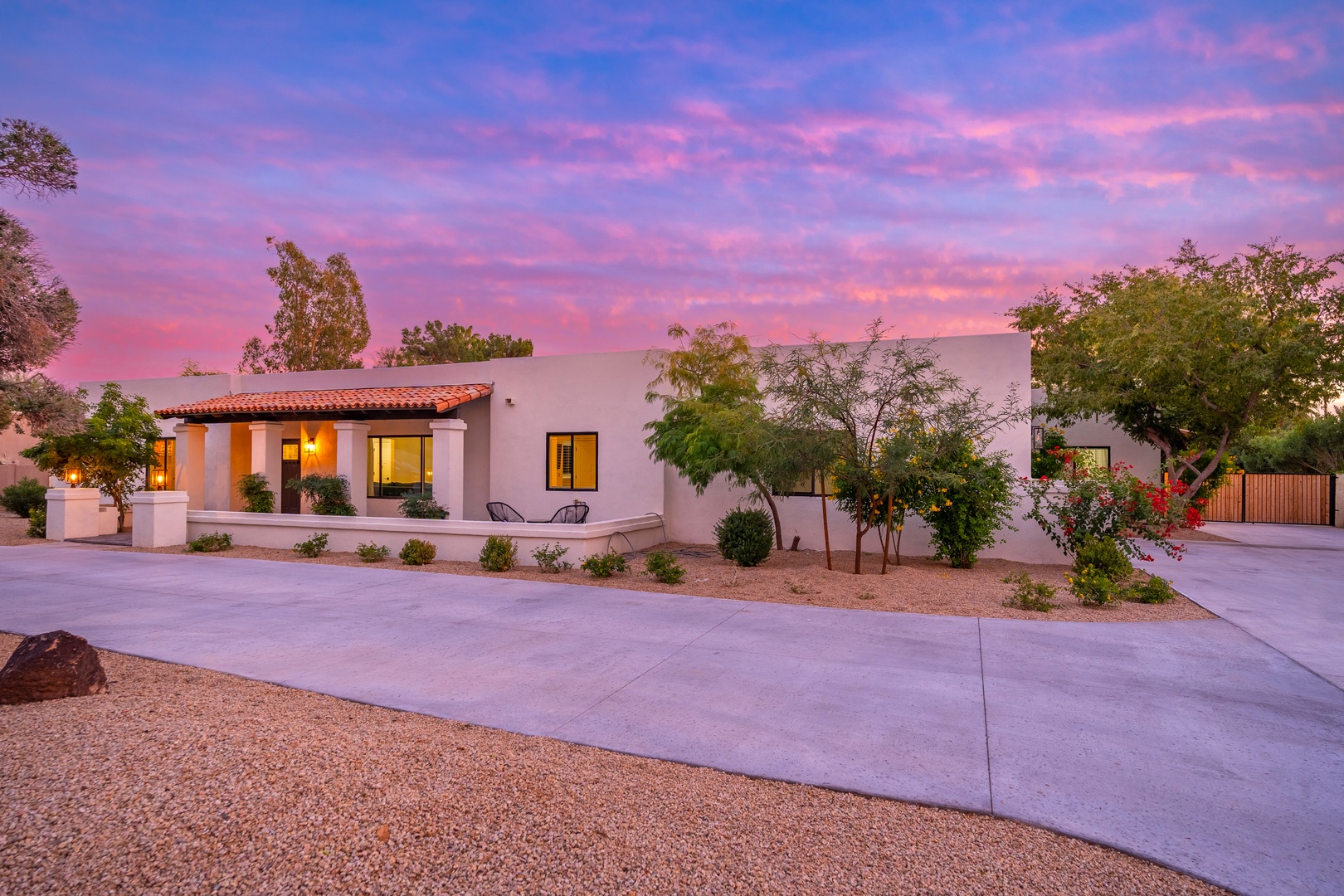 Striking desert retreat with Spanish-style tile roof and mature landscaping, framed by a vibrant pink and purple sunset sky, offering a welcoming Southwest atmosphere upon arrival.