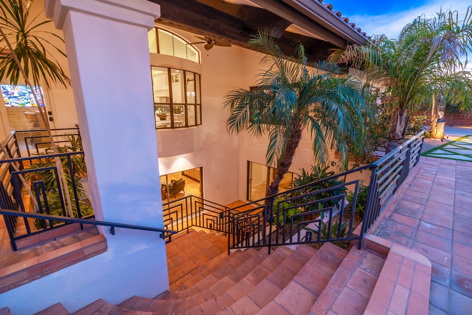 A stunning two-story villa entrance showcasing elegant Spanish-style architecture with terracotta tile steps, lush palm trees, and an inviting courtyard bathed in warm evening light under clear blue skies.