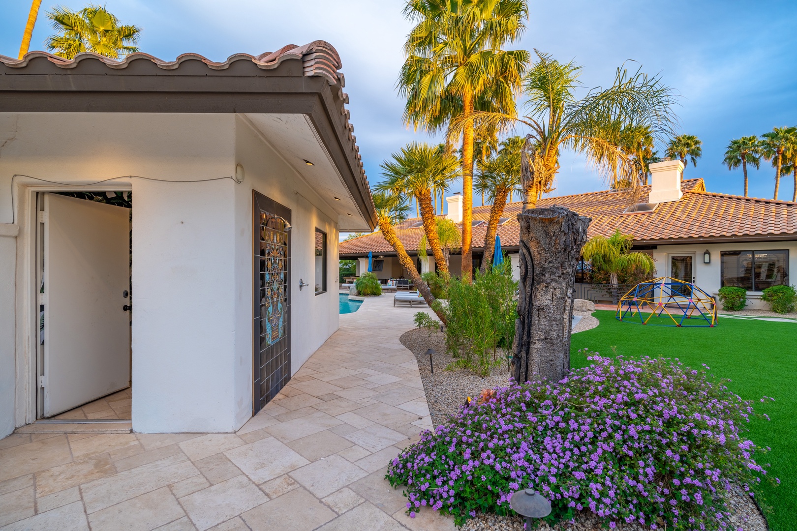A resort-style property with terracotta roofs, towering palms, and vibrant purple blooms welcomes guests to a lush desert retreat with pool and playground.