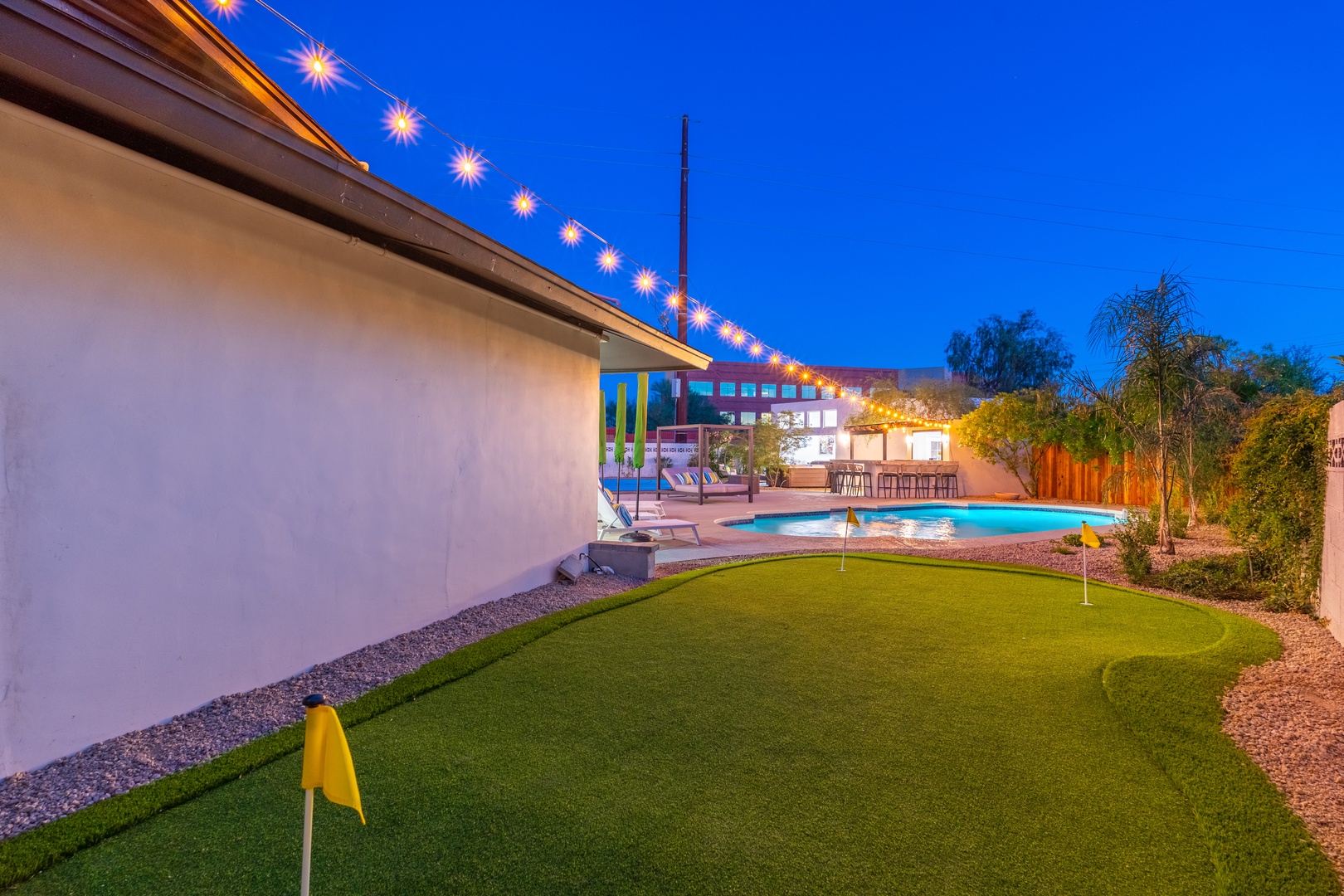 A twilight view captures the property's backyard oasis, featuring a private putting green, sparkling pool, colorful entertainment area, and festive string lighting beneath a vibrant desert sky.
