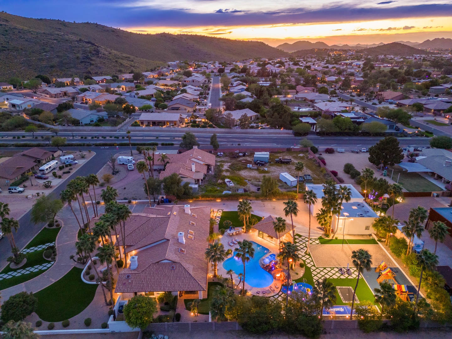 Aerial view of a spacious vacation estate nestled in a scenic neighborhood, featuring a sparkling pool and lush palm trees, captured at golden hour with mountains in the distance.