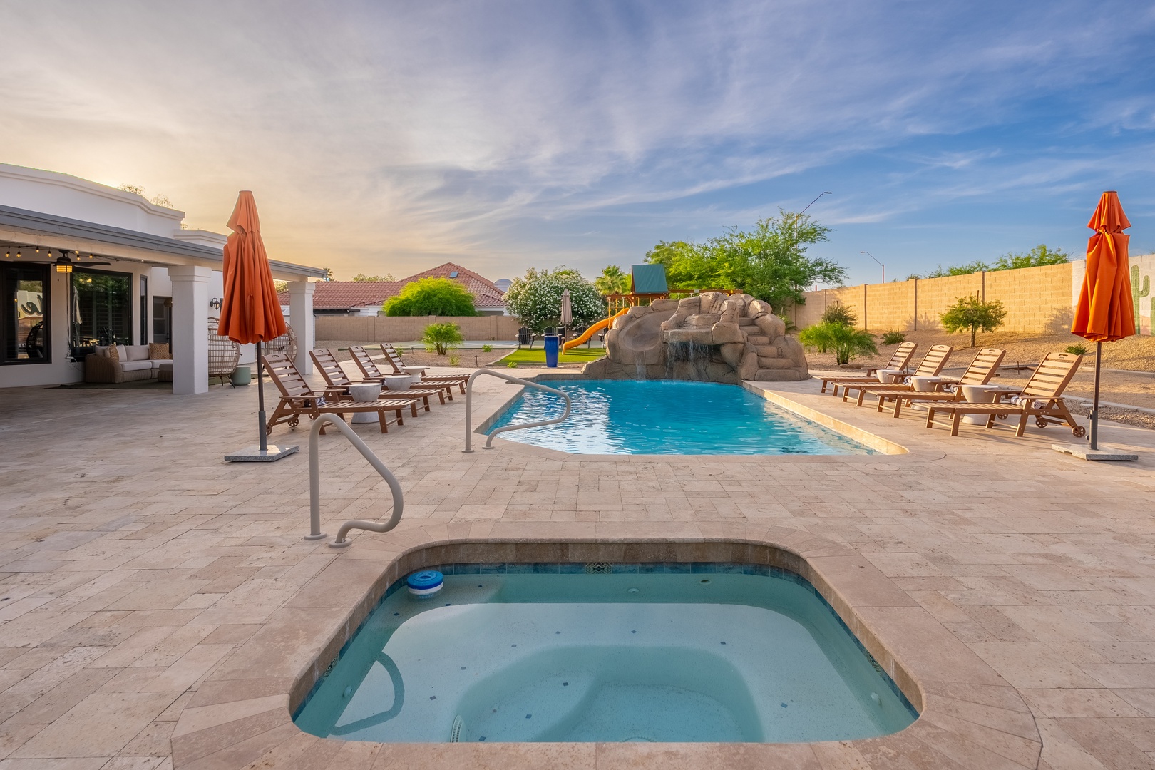 Resort-style backyard featuring a sparkling swimming pool, cascading waterfall, bubbling hot tub, and ample lounge seating under desert skies.