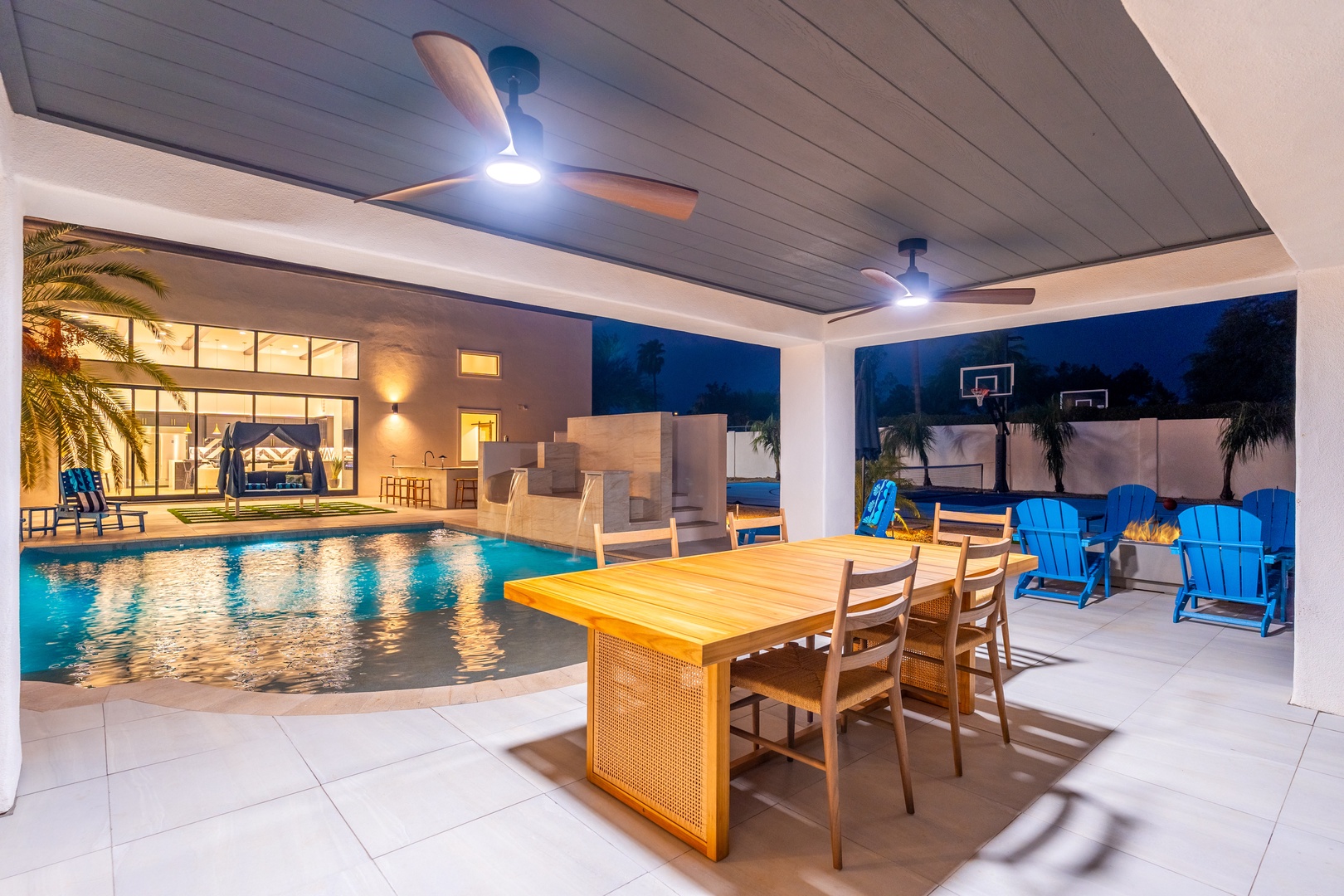 Your private covered patio features poolside dining and vibrant blue Adirondack chairs, perfect for evening gatherings under ceiling fans with pool views and basketball fun nearby.