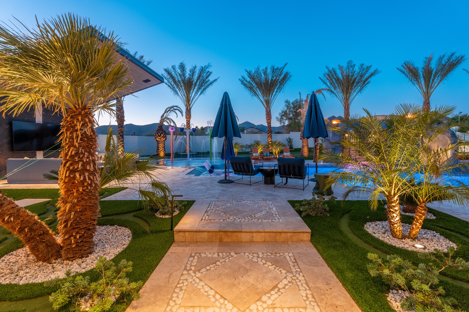 The property features a stunning outdoor oasis with manicured gardens, illuminated palm trees, and a designer pool area, set against a backdrop of distant mountains and open skies.