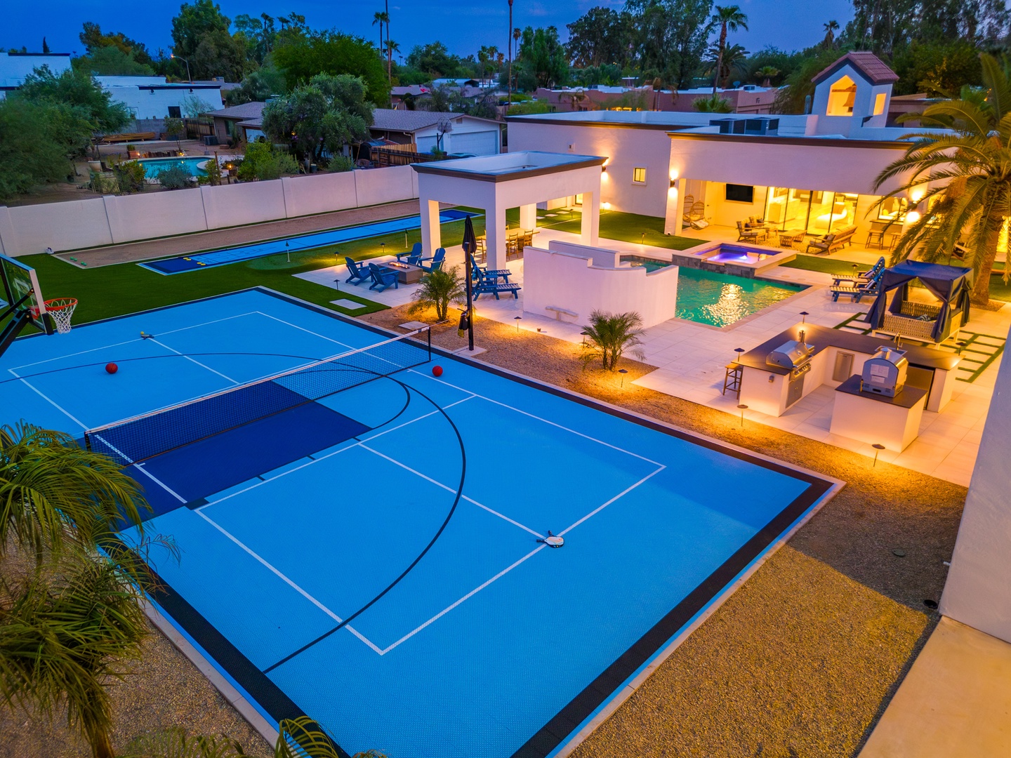 This modern retreat features a private sports court, sparkling pool with hot tub, and multiple outdoor lounging areas—all illuminated for evening enjoyment beneath palm trees and warm desert skies.
