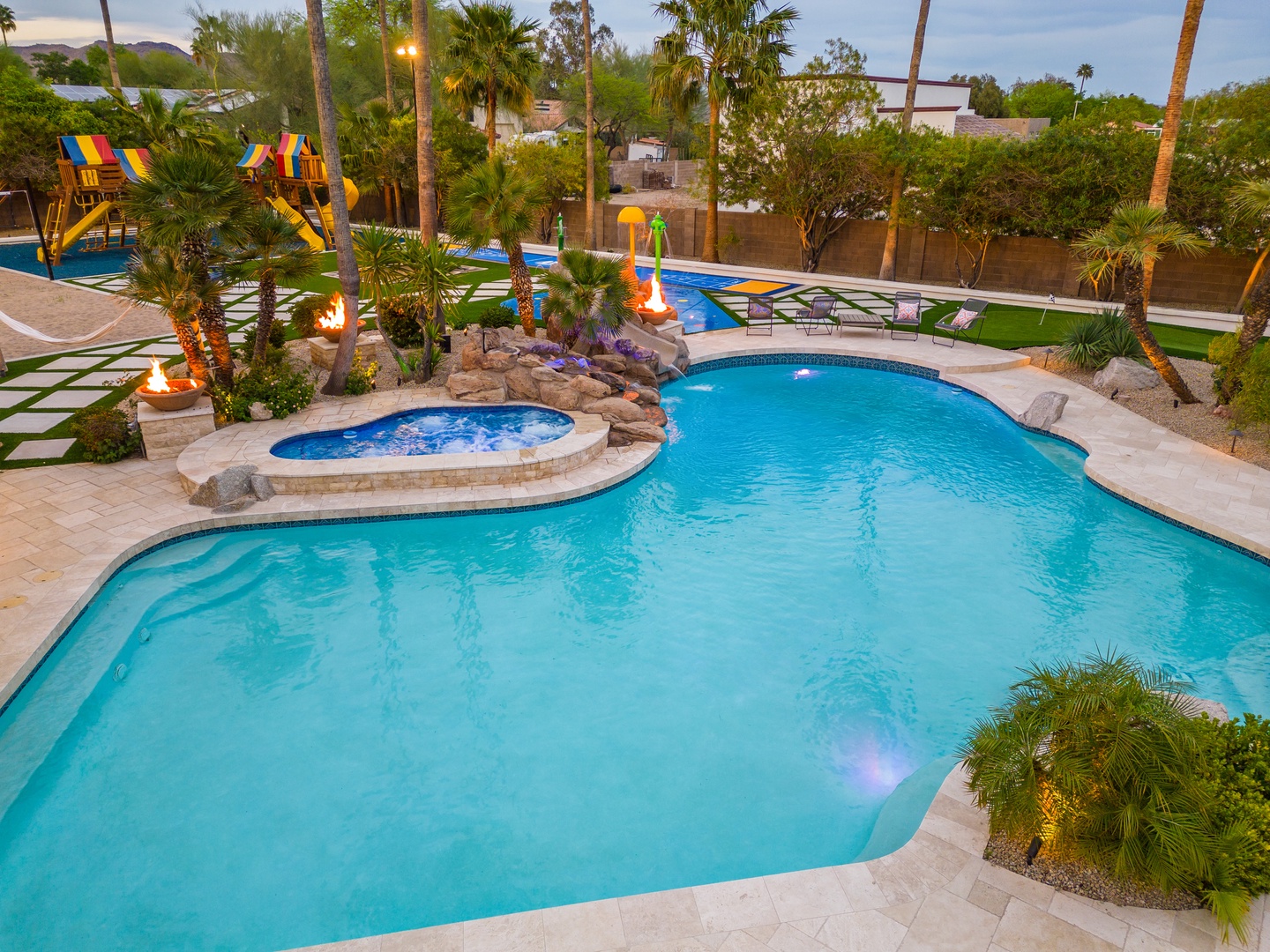 An expansive backyard oasis featuring a freeform pool with integrated hot tub, palm trees, playground, and spacious lawn areas—perfect for family gatherings.