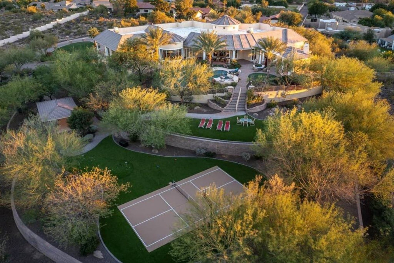 Sprawling luxury estate set within landscaped grounds featuring a tennis court, swimming pool, and lush desert foliage in a peaceful residential neighborhood.