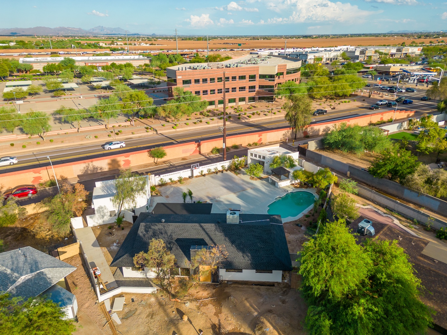 Aerial view shows the property positioned in a residential area with nearby commercial buildings, tree-lined streets, and a private pool. The location offers convenient access to surrounding amenities and services.