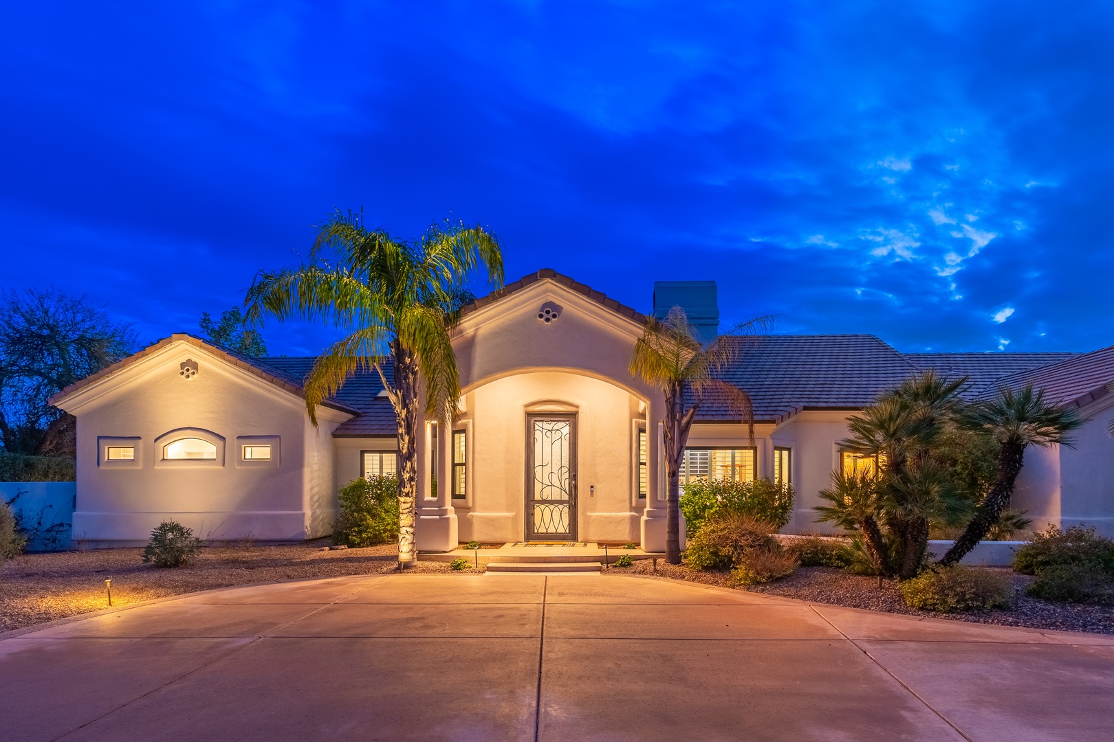 A welcoming single-story home with elegant architecture, warmly lit under a twilight sky, surrounded by lush palm trees and desert landscaping in a peaceful residential setting.