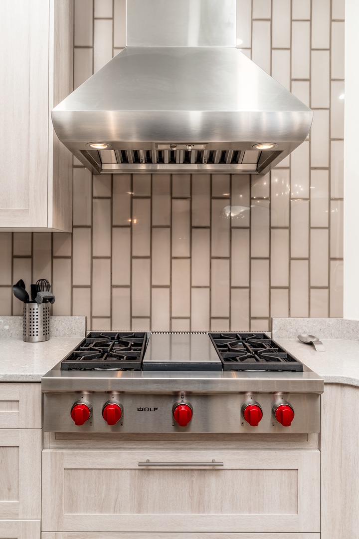 Cook your favorite meals on a high-end gas range with bold red knobs, complemented by a professional-grade stainless steel hood and elegant white countertops.
